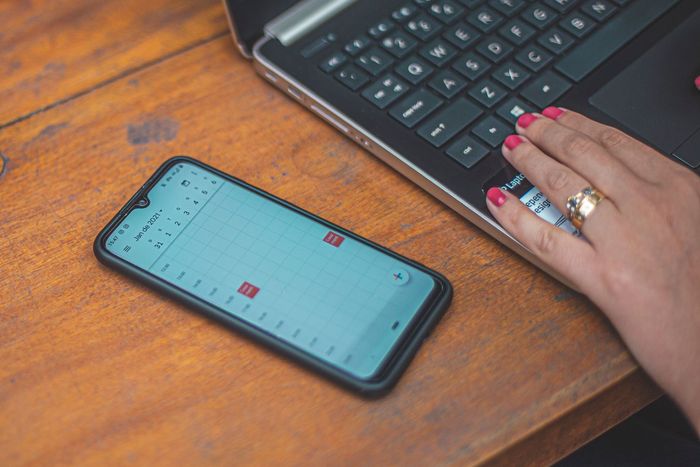 A mobile phone showing a month calendar on a desk, next to a laptop being used.